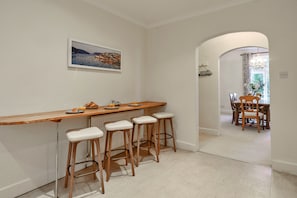 A kitchen with a wooden bar, four stools, a dining table with chairs, and a painting on the wall. - Grand Salcombe Retreat (Salcombe)