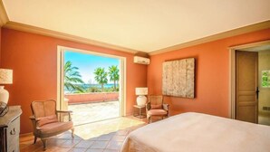 A hotel room with a bed, two chairs, a lamp, and a large window with a view of palm trees and the ocean. - Villa 'Maison Rouge - Esprit Du Sud' with Sea View, Private Pool and Wi-Fi (Sainte-Maxime)