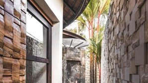 A narrow outdoor corridor with a stone wall on one side and a wooden wall on the other, leading to a covered area with a stone fireplace. - Sandi Kala Villas - Two-Bedroom Villa (Badung Regency)