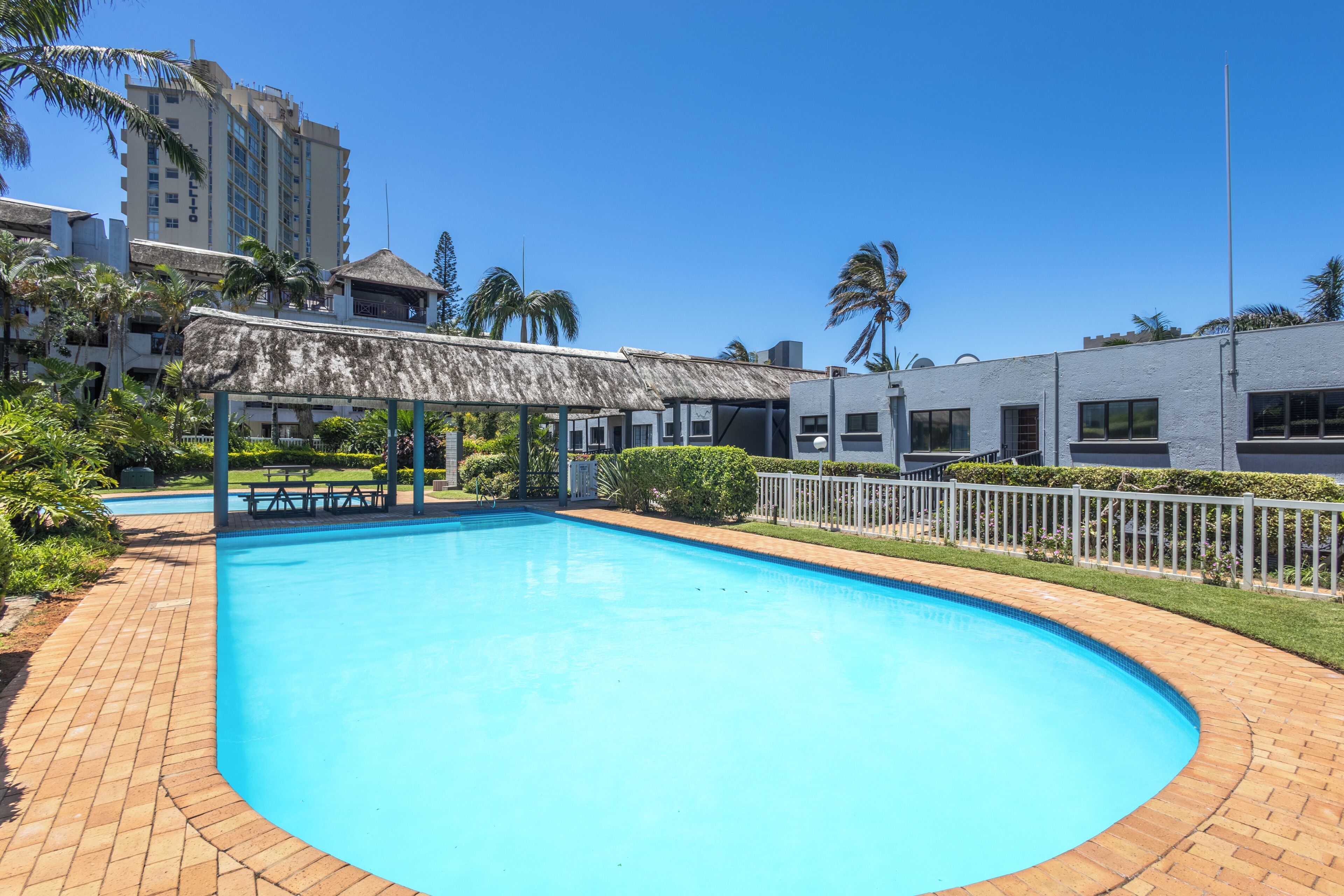 A swimming pool with a thatched-roof pavilion, surrounded by a brick walkway and palm trees.