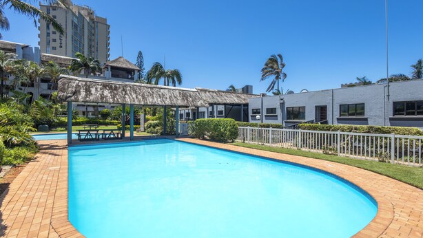 A swimming pool with a thatched-roof pavilion, surrounded by a brick walkway and palm trees.
