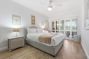 A bedroom with a bed, nightstand, lamp, ceiling fan, and a view through a window with blinds. - Paradise Beach Apartment | Pool | Ballito (Dolphin Coast)