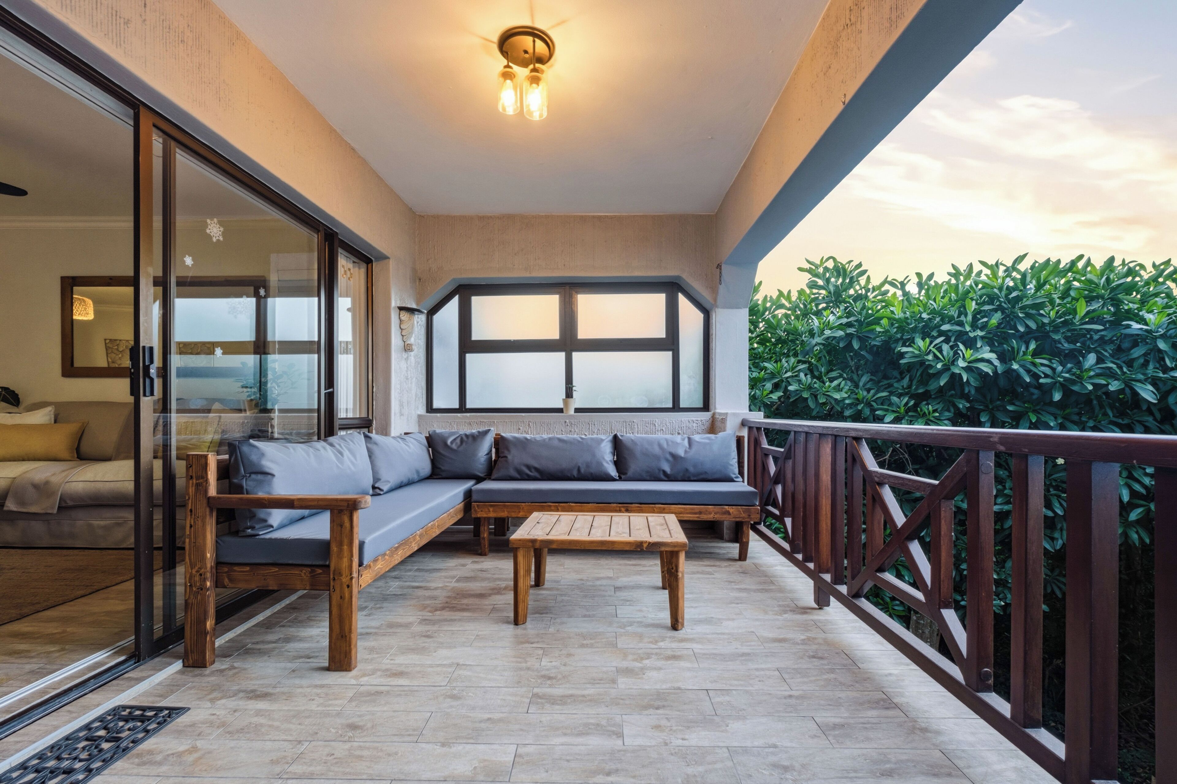 A balcony with a wooden bench and a sofa, a glass door leading to a room, and a view of greenery.