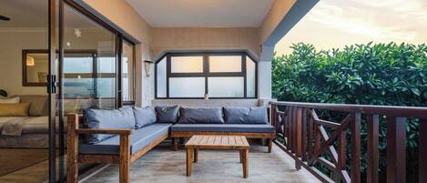 A balcony with a wooden bench and a sofa, a glass door leading to a room, and a view of greenery.