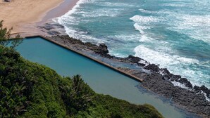 A coastal landscape with a rocky shoreline, a clear blue pool, and dense green vegetation.