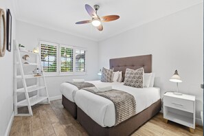 A bedroom with a bed, bedside tables, a ceiling fan, a window with blinds, and a white wardrobe. - Paradise Beach Apartment | Pool | Ballito (Dolphin Coast)