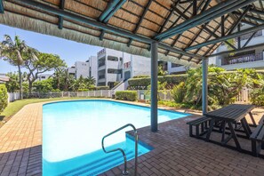 A swimming pool with a thatched roof structure, surrounded by a brick patio and wooden picnic tables. - Paradise Beach Apartment | Pool | Ballito (Dolphin Coast)