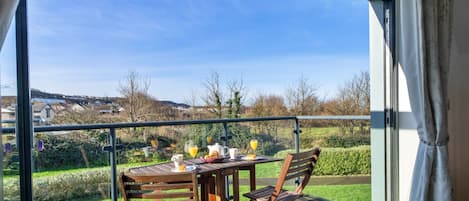 A balcony with a table set for breakfast, including croissants and orange juice, overlooking a green landscape.