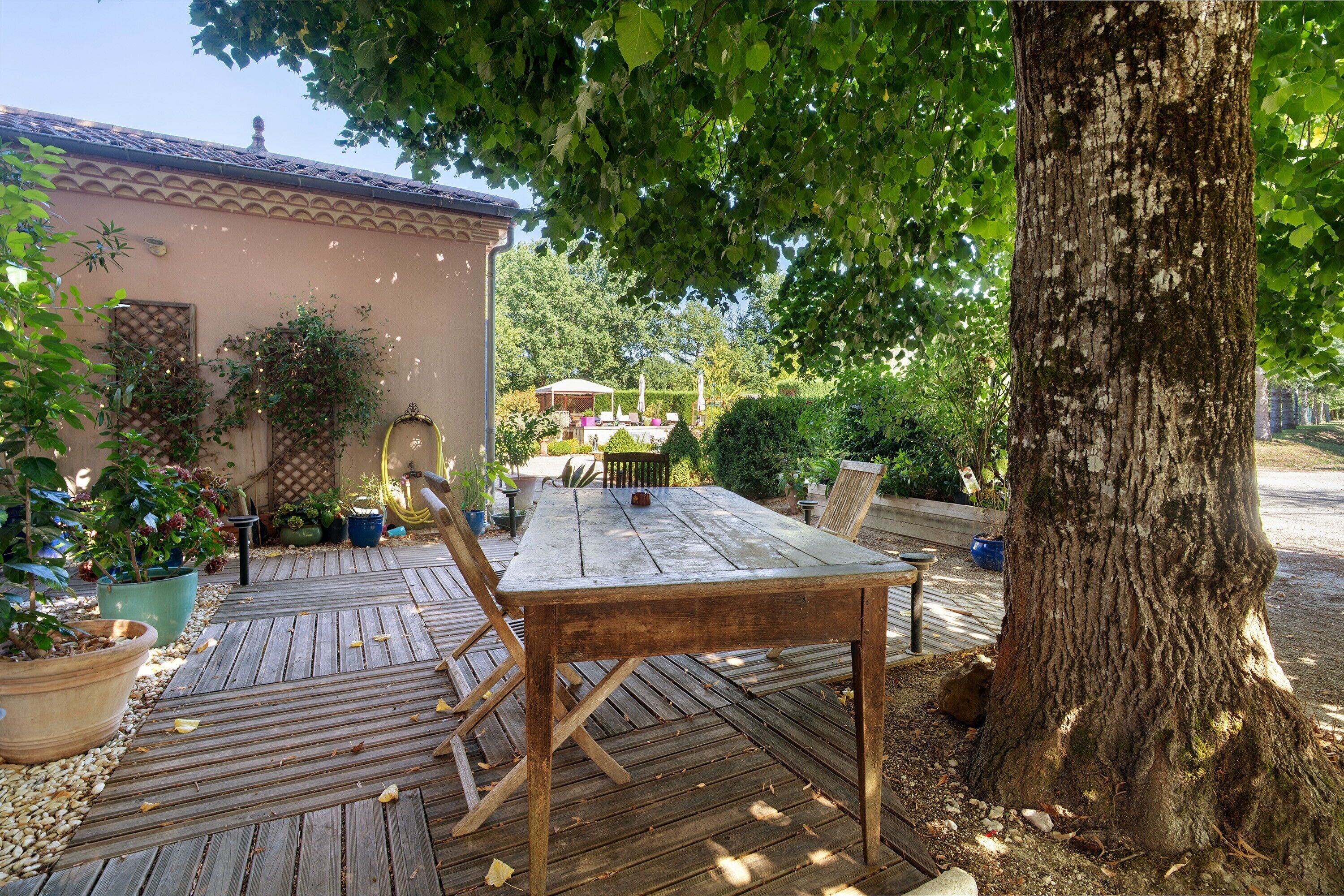 Une terrasse extérieure en bois, avec une table et des chaises, entourée de plantes en pot et d’un grand arbre.