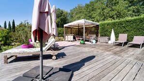 Una terraza de madera con mobiliario exterior, incluyendo sillas y una mesa, y un área cubierta con una mesa y sillas.