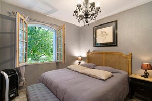 A bedroom with a bed, a window, a chandelier, a bedside table, and a framed artwork.