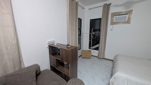 Une chambre d’hôtel avec un lit, une table de chevet en bois, un bureau et une chaise. Il y a une fenêtre avec des rideaux, une porte et un petit appareil de ventilation fixé au mur.