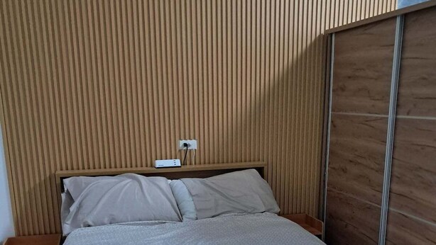 A modern bedroom with a wooden headboard, a bed with a patterned bedspread, a bedside table, and a power outlet. - City View Studio - Pool · Gym - Caesar Residence (Lapu-Lapu)