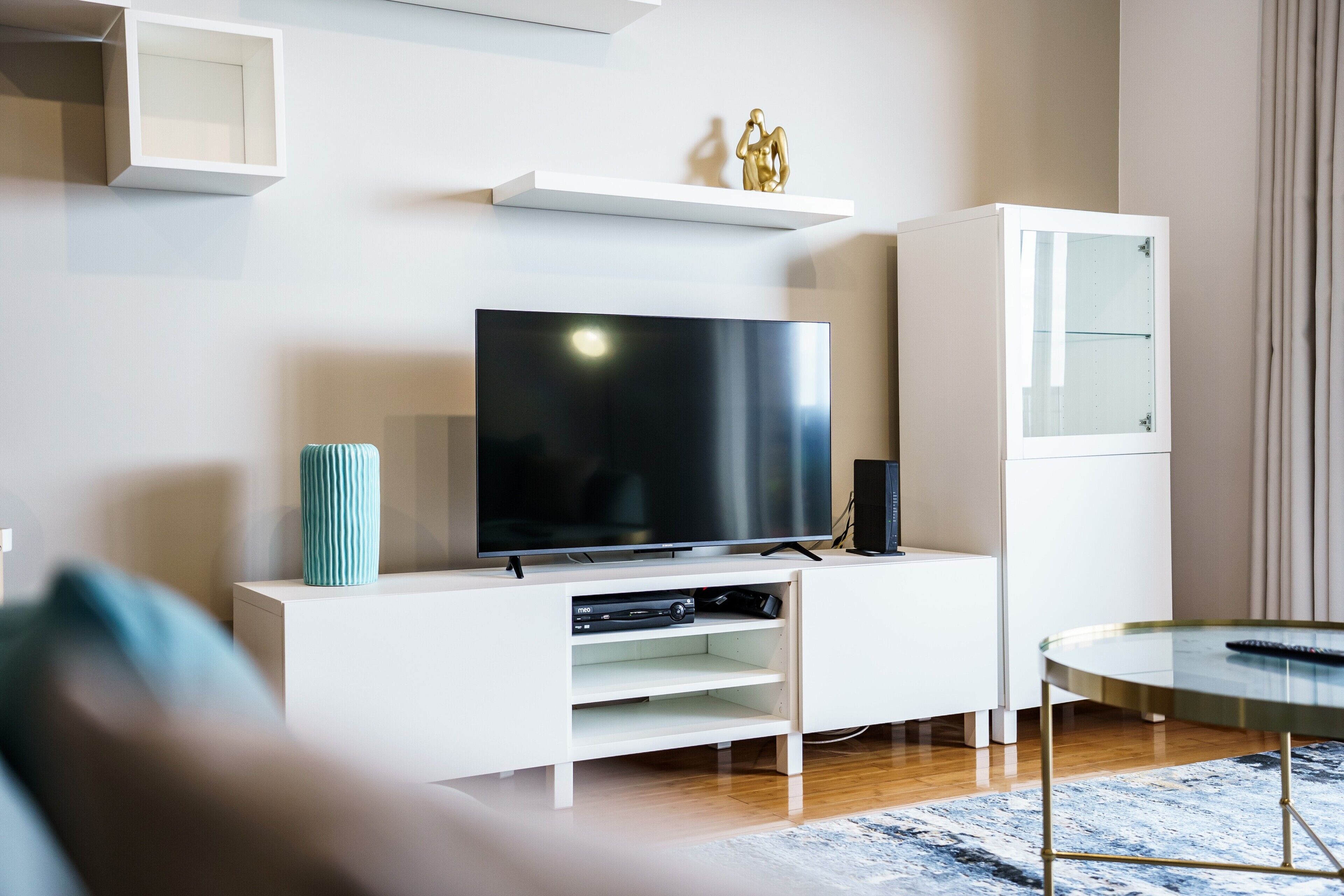 Un salon moderne avec un téléviseur à écran plat posé sur un meuble de télévision blanc, une vitrine et une petite table ronde sur laquelle se trouve une télécommande.