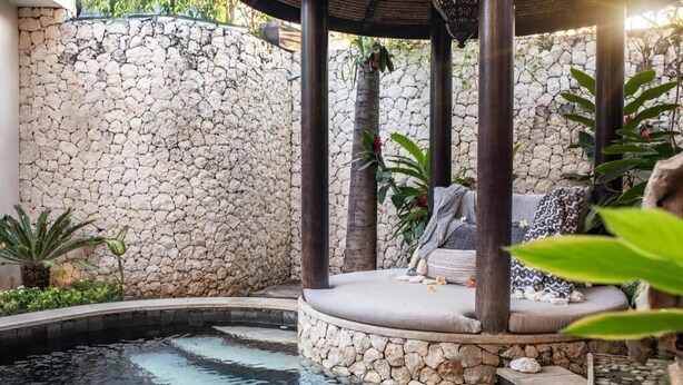 Um muro de pedra, uma área de estar circular com almofadas e uma pequena piscina.