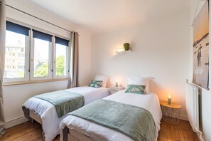 Ein Hotelzimmer mit zwei Betten, einem Nachttisch mit Lampe, einer Wandleuchte und einem Fenster mit Vorhängen.