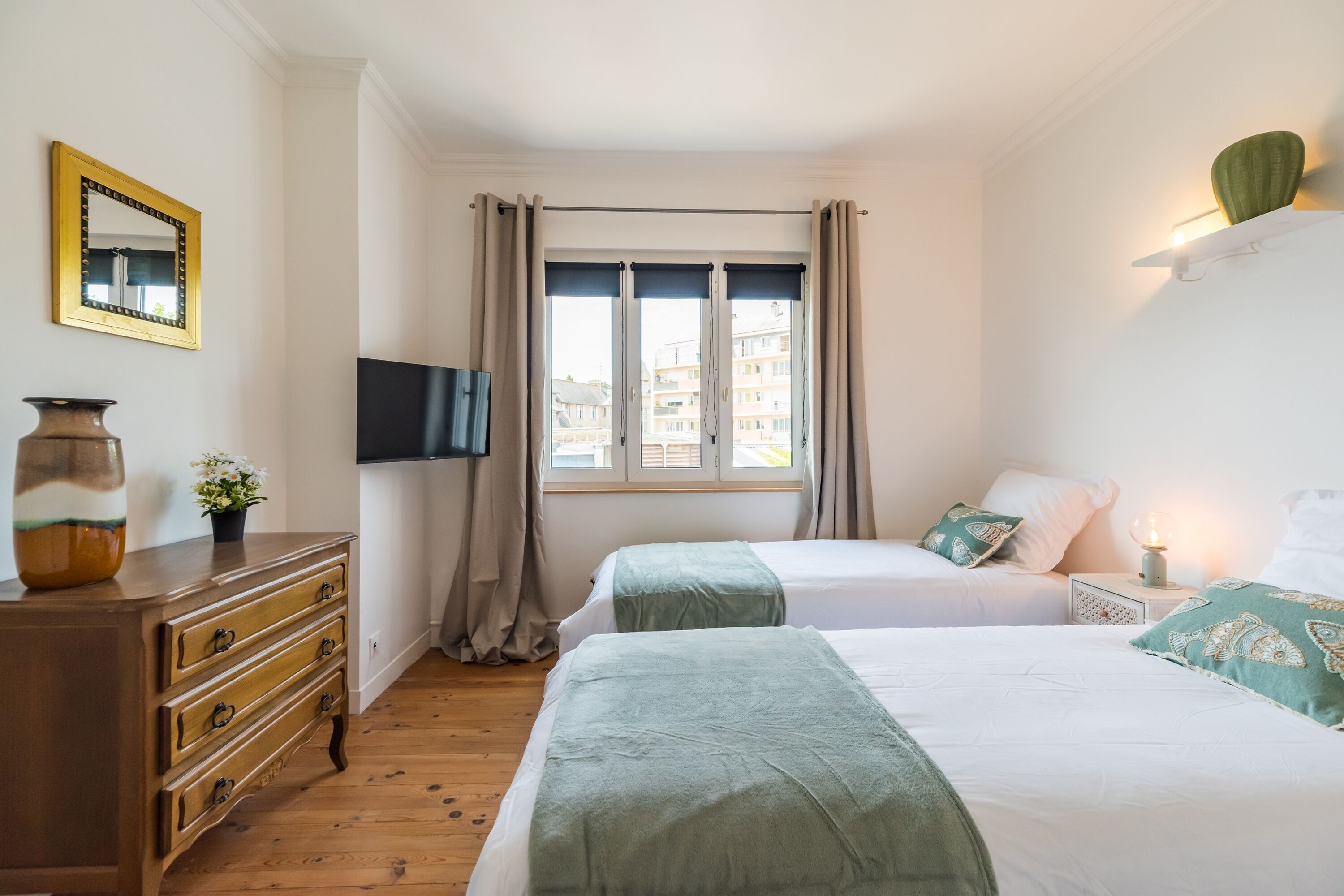 A hotel room with two beds, a dresser, a flat-screen TV, and a window with curtains.