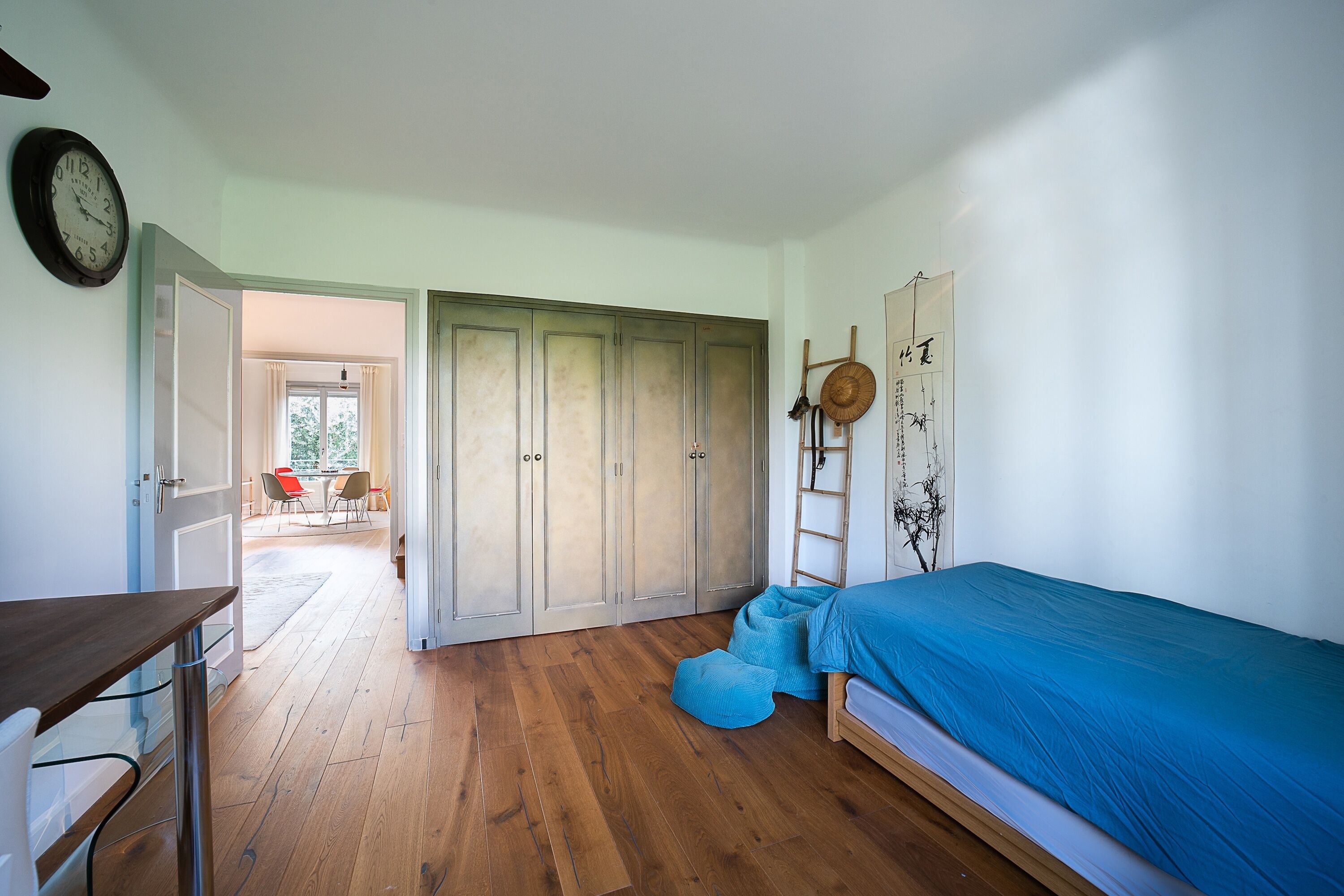 A bedroom with a wooden floor, a bed with a blue cover, a wardrobe, a desk, and a clock on the wall.