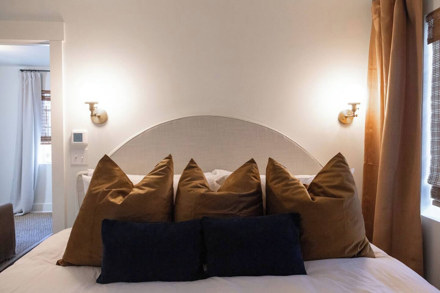 A hotel room with a large bed, multiple pillows, a headboard, wall sconces, and a window with curtains.