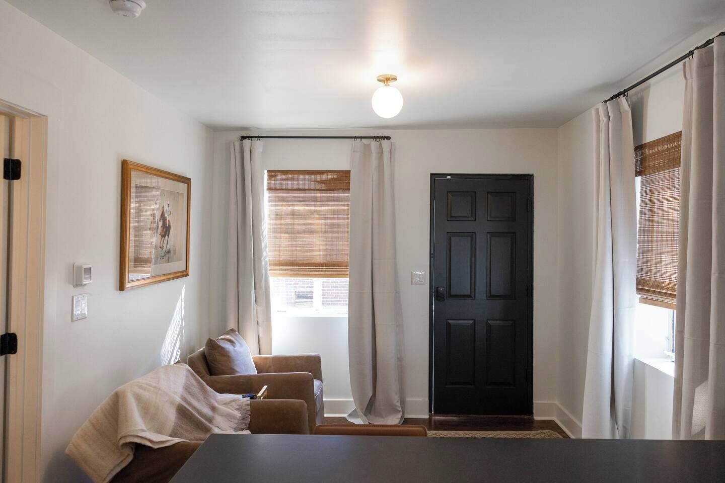 A room with a dark door, a window with blinds, a framed picture on the wall, and a sofa with a blanket.