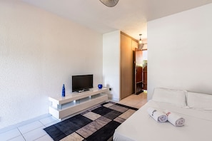 A modern bedroom with a bed, a TV on a white stand, a black and white rug, and a wooden door. - La Plage d'Irène (Appartement n°36) - Avec balcon (San-Nicolao)
