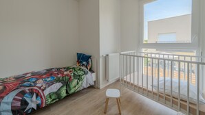 Ein Schlafzimmer mit einem Einzelbett, einem kleinen weißen Hocker, einem weißen Heizkörper und einem Balkon mit weißer Brüstung.