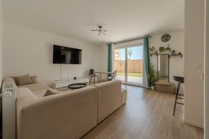 A modern living room with a flat-screen TV, a sofa, a dining table, and a sliding glass door leading to an outdoor area. - Le Cocon des Yvelines - Jolie maison avec jardin (Aubergenville)