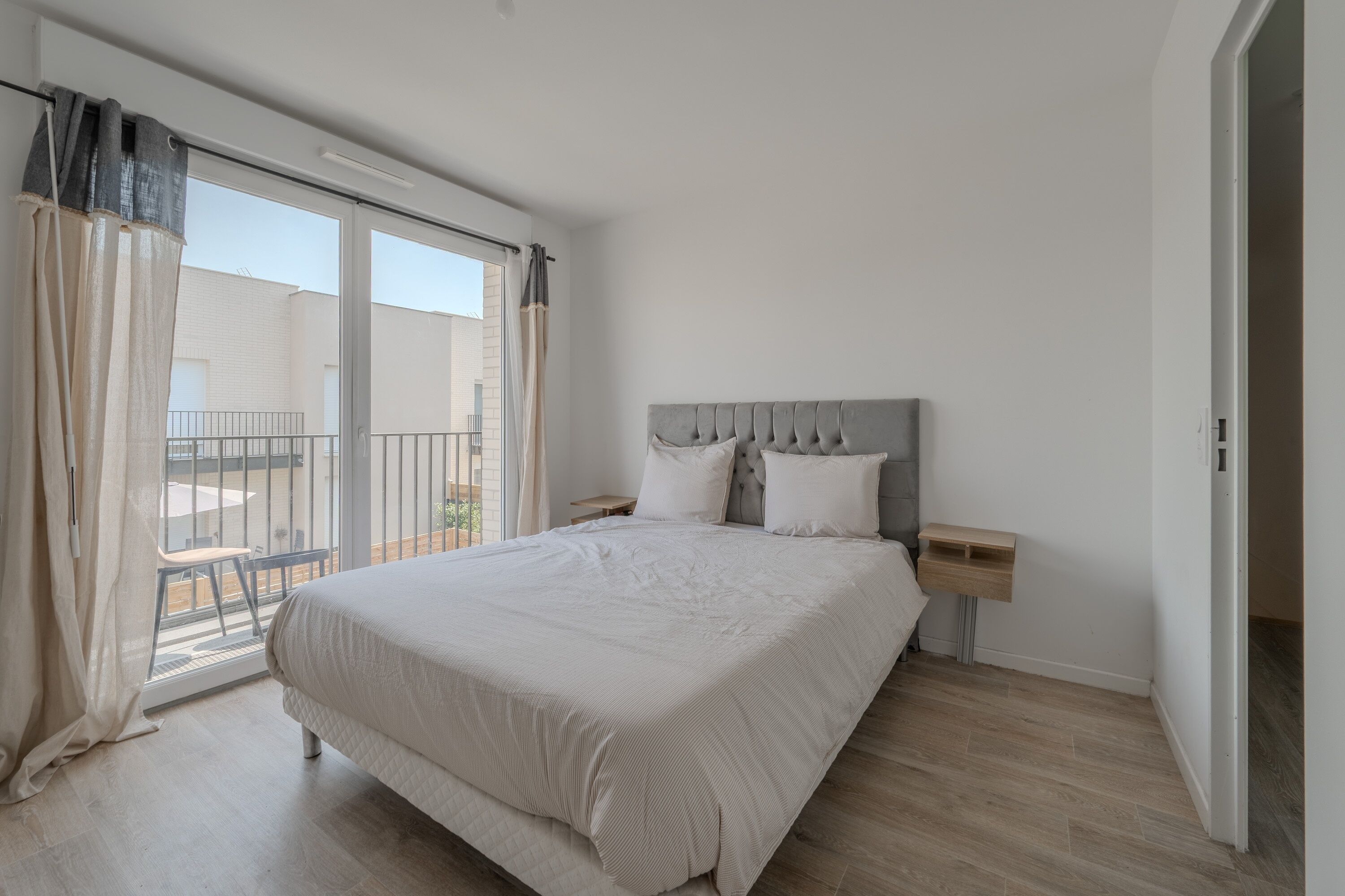 A bedroom with a large bed, a nightstand, a window with curtains, and a view of the outside.