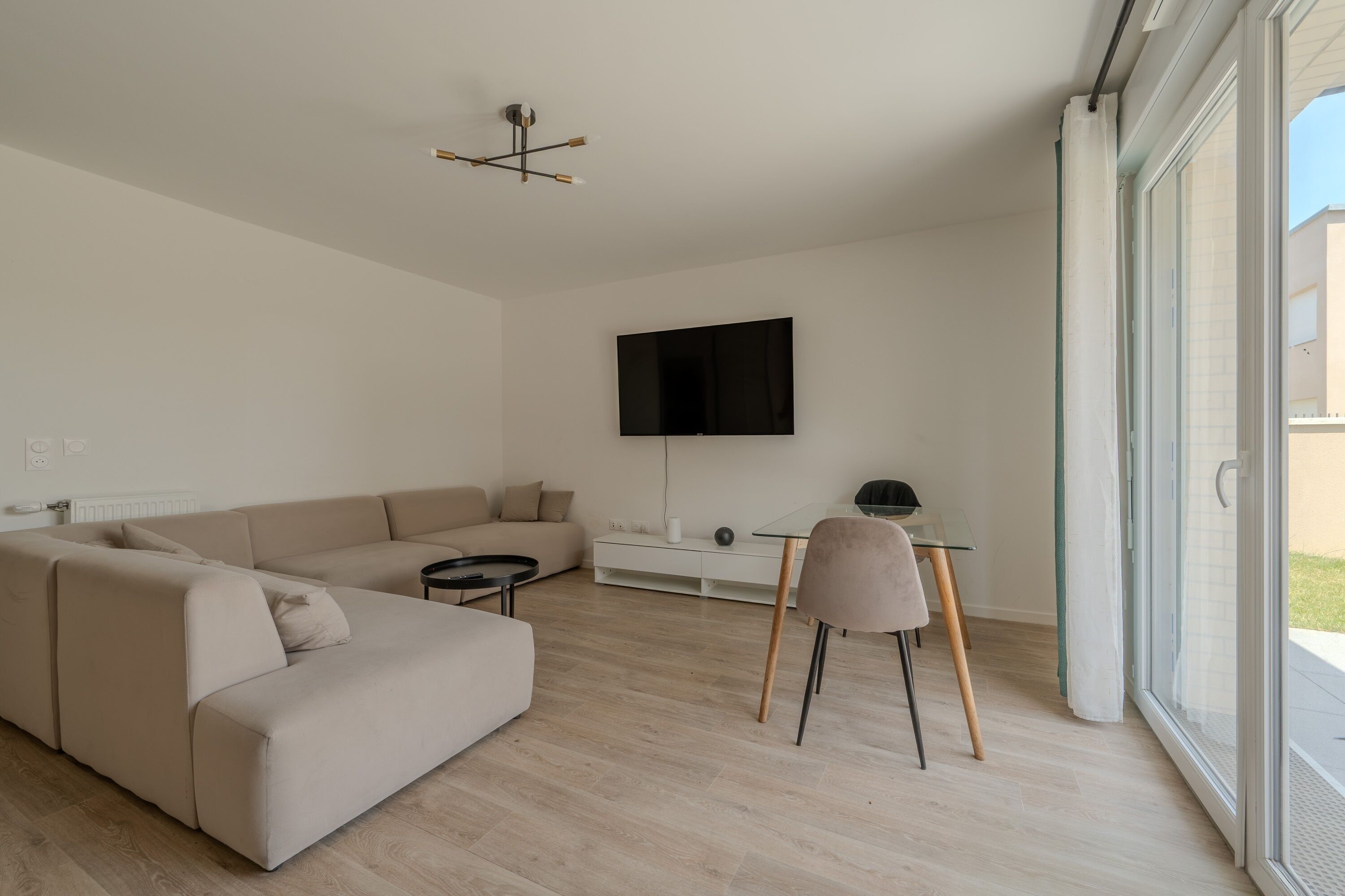 A modern living room with a sofa, a coffee table, a TV mounted on the wall, and a glass door leading to an outdoor area.