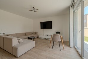 A modern living room with a sofa, a coffee table, a TV mounted on the wall, and a glass door leading to an outdoor area. - Le Cocon des Yvelines - Jolie maison avec jardin (Aubergenville)