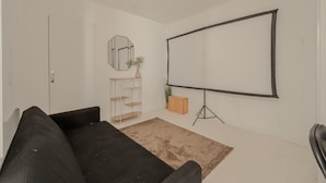 Ein minimalistisches Wohnzimmer mit einer schwarzen Couch, einer weißen Wand, einer großen, leeren Projektionsleinwand und einem kleinen Holzregal mit einer Pflanze.