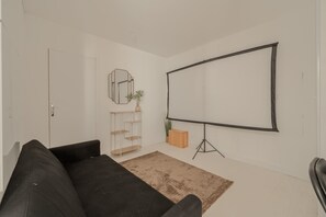 A minimalist living room with a black sofa, a white wall, a large blank projection screen, and a small wooden shelf with a plant. - Le Cocon des Yvelines - Jolie maison avec jardin (Aubergenville)