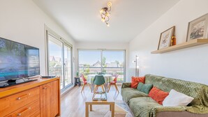 A modern living room with a flat-screen TV, wooden cabinet, green sofa, and a dining area with a table and chairs. - La Plage - À moins de 100 m de la plage (Courseulles-sur-Mer)