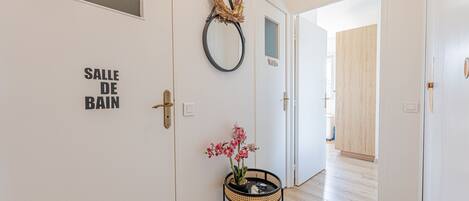 Un couloir avec une porte sur laquelle est inscrit « SALLE DE BAIN » et une petite table sur laquelle est posée une orchidée en pot.