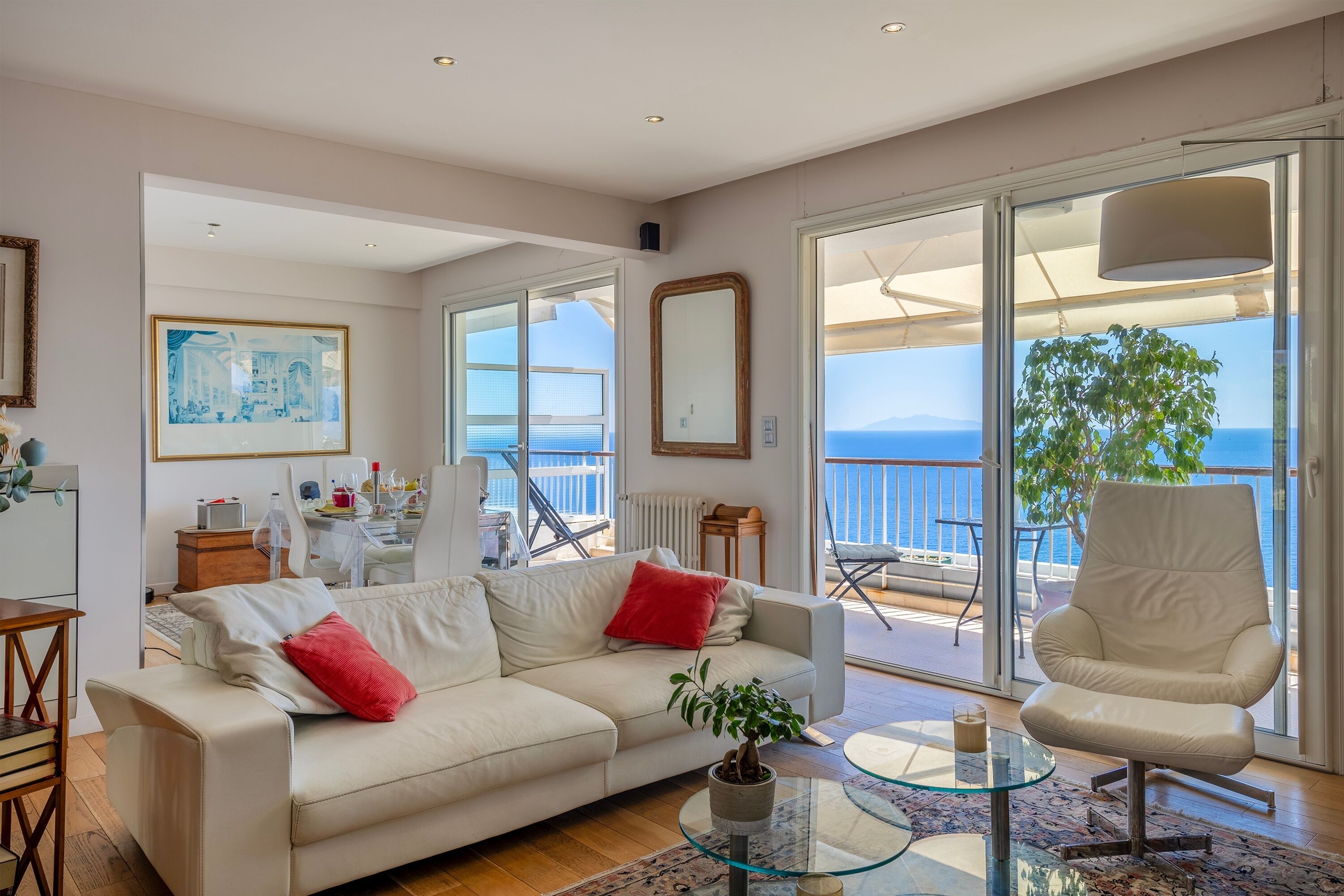 Un salon spacieux avec un canapé blanc, des coussins rouges, une table basse en verre et une vue sur la mer grâce à de grandes fenêtres.