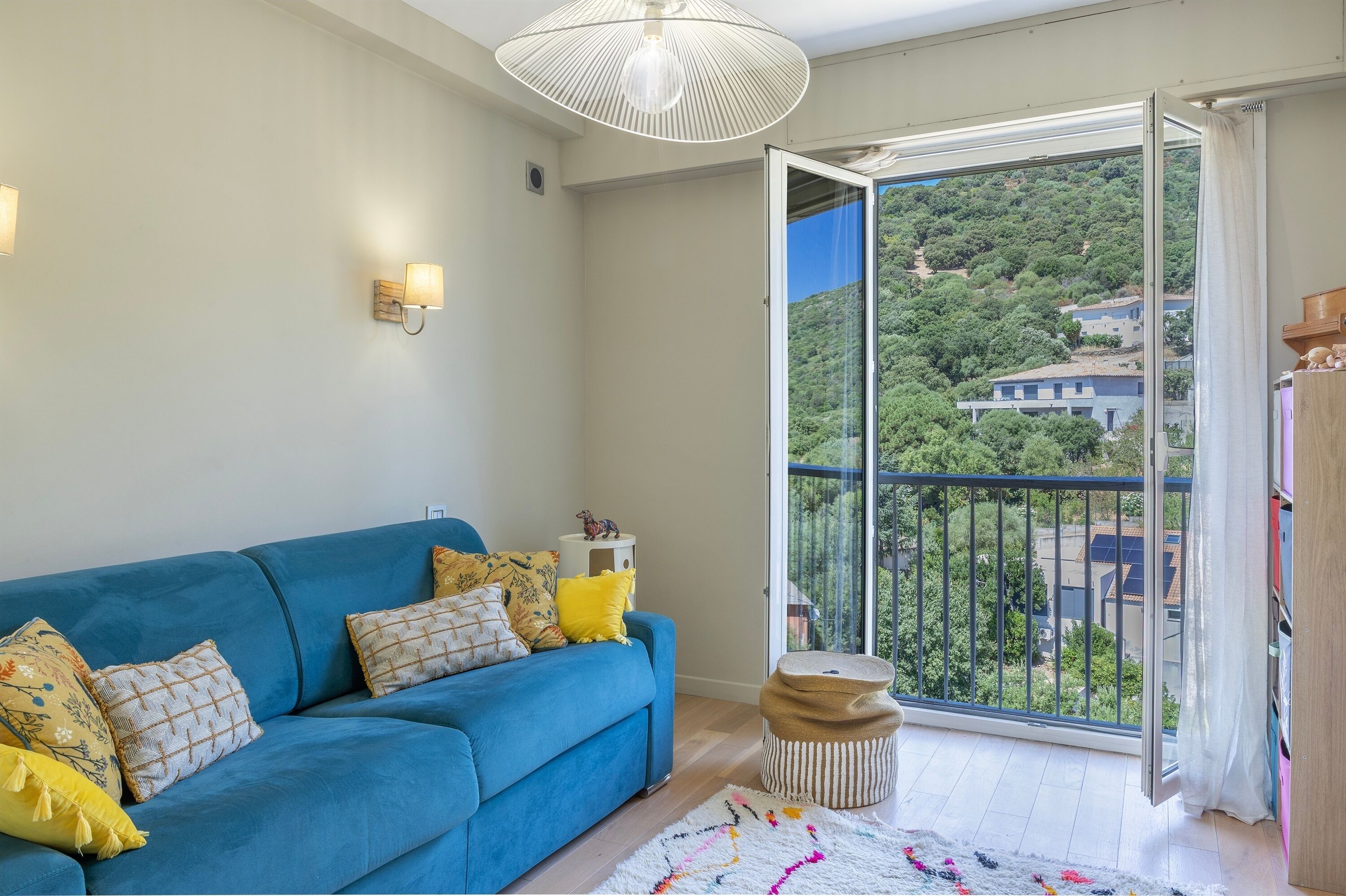 Un salon avec un canapé bleu, un petit pouf rond, une lampe blanche et une vue sur un paysage verdoyant, visible à travers une porte-fenêtre donnant sur un balcon.