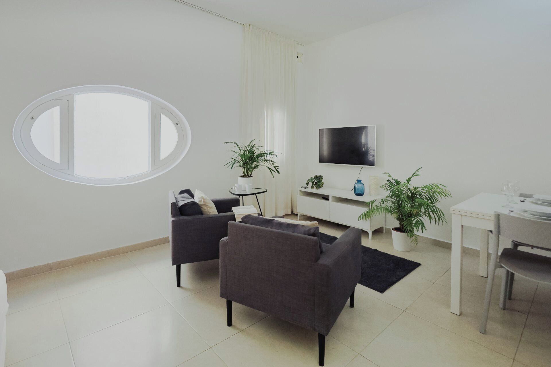 Un salon moderne avec une fenêtre ronde, des canapés gris, un meuble de télévision blanc et une table à manger.