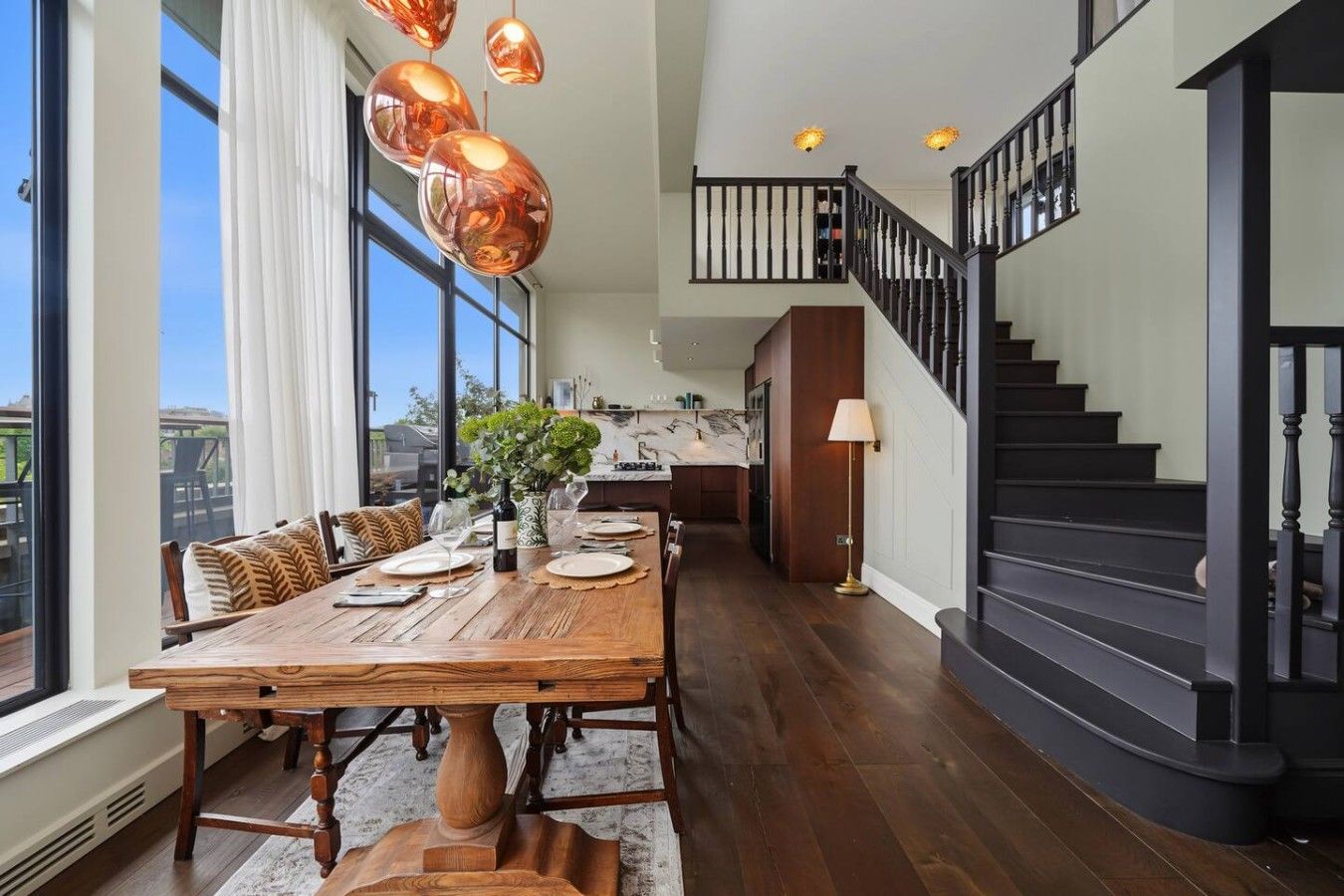 A dining area with a wooden table set for six, a kitchen in the background, and a staircase with black railings.