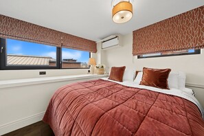 Ein Schlafzimmer mit einem großen Bett, zwei Fenstern mit gemusterten Vorhängen, einer Klimaanlage und Blick auf Gebäude.