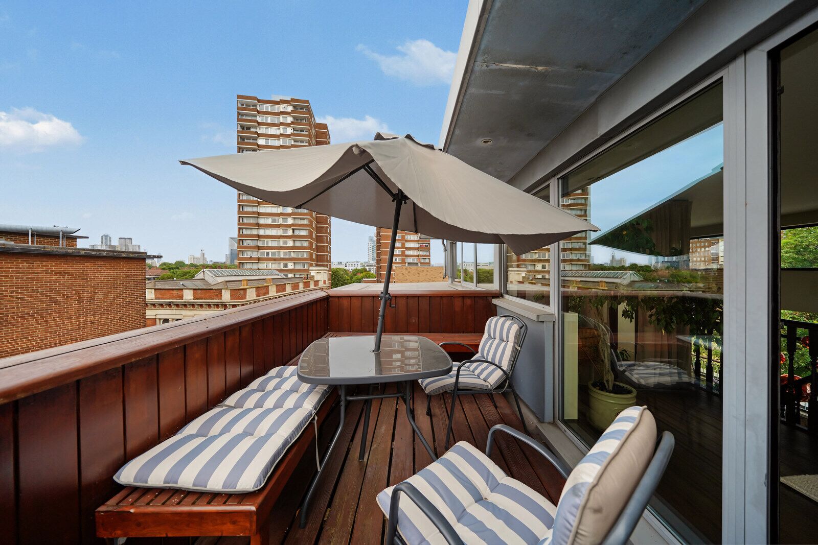 Eine Dachterrasse mit Tisch und Stühlen, einem großen Sonnenschirm und Blick auf umliegende Gebäude.