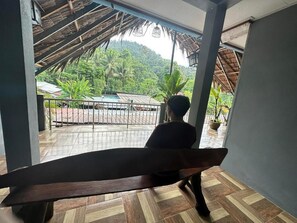 En person sitter på en bänk inne i en byggnad och har utsikt över en balkong med en vacker vy över ett frodigt, grönt landskap och en byggnad med ett halmtak.