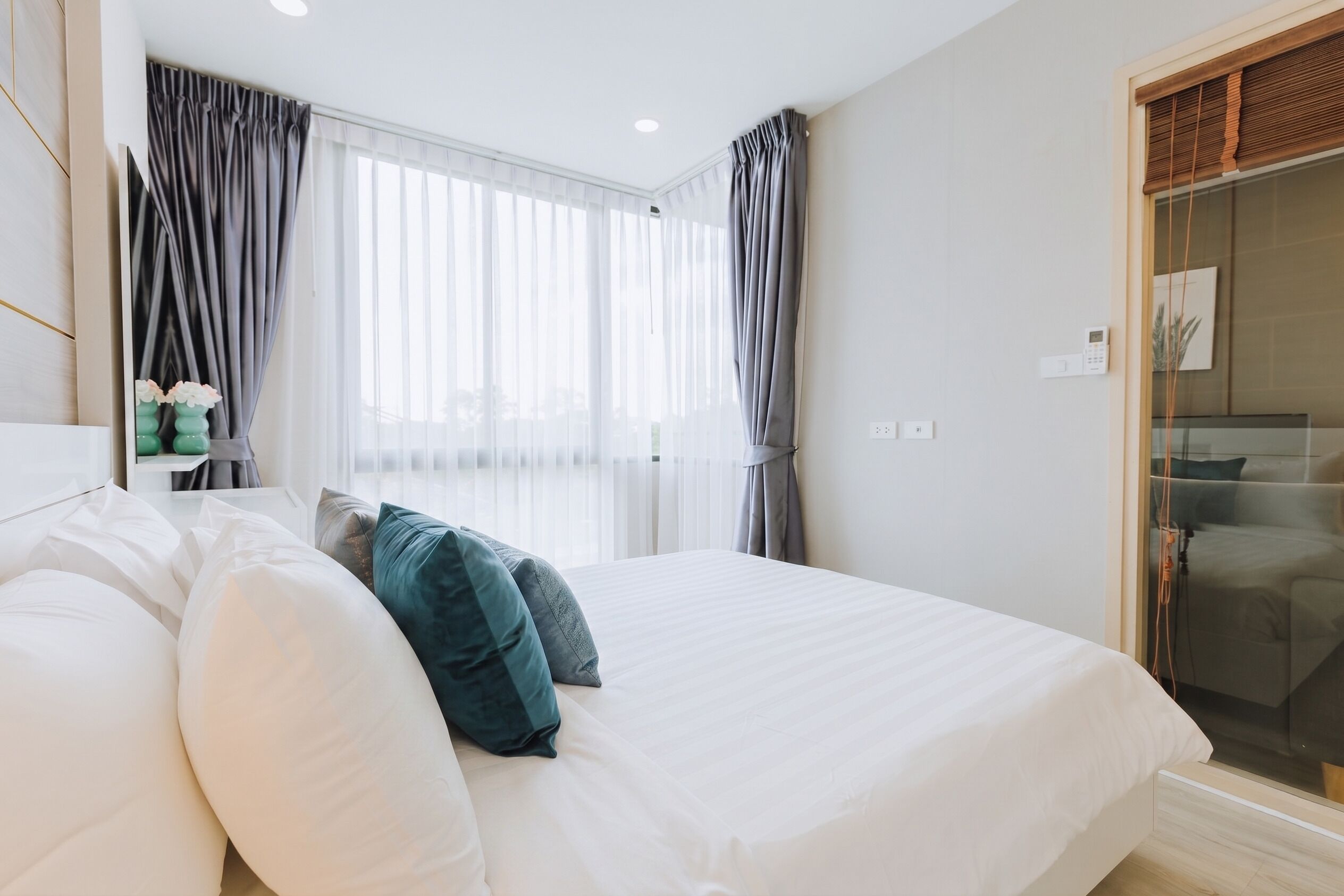 Une chambre avec un grand lit, des rideaux gris, une table de chevet avec un vase, et une vue à travers une porte vitrée.