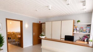 A modern reception area with a wooden counter, a seating area with a sofa and a coffee table, and a dining area visible through an open door. - Hotel Esingerhof by Alesta (Tornesch)