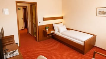 A hotel room with a bed, desk, chair, and a small bedside table with a lamp.