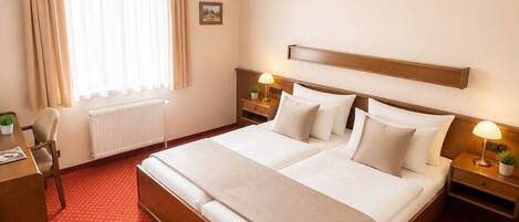 Quarto de hotel com uma cama grande, duas mesas de cabeceira, uma escrivaninha, uma cadeira, um radiador e uma janela com cortinas.