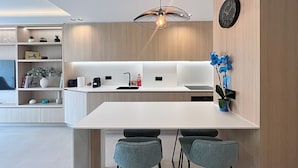 Eine moderne Küche mit weißer Arbeitsplatte, zwei Barhockern, einer hängenden Leuchte und einer Uhr an der Wand.