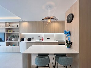 Eine moderne Küche mit weißer Arbeitsplatte, zwei Barhockern, einer hängenden Leuchte und einer Uhr an der Wand.