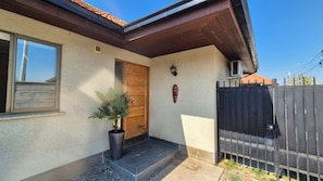 A house with a wooden door, a potted plant, a small wall-mounted light, and a black metal fence.
