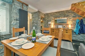 A dining area with a wooden table set for four, a bottle of wine, and a stone wall backdrop. - Charming Snowdonia Slate Cottage (Blaenau Ffestiniog)