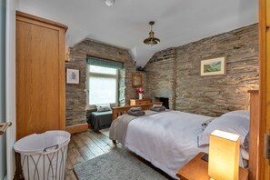 Un dormitorio con paredes de piedra, un armario de madera, una cama con almohadas, una mesita de noche con lámpara y una ventana con vistas.